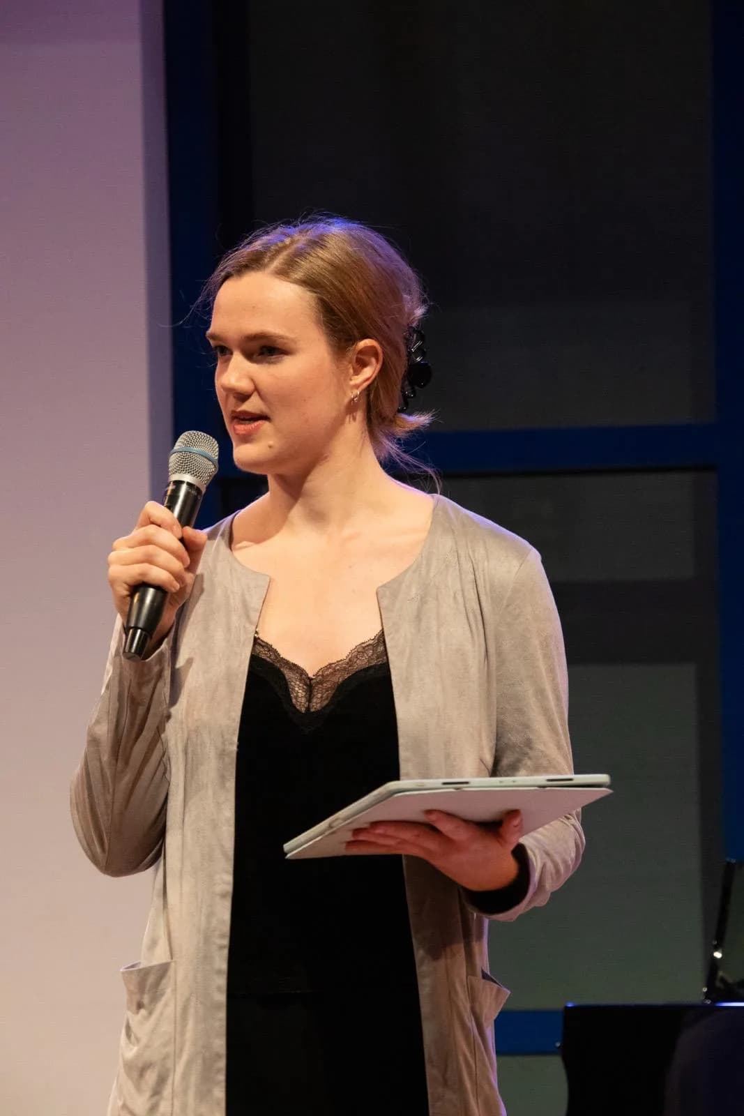 Annika Spegg im grauen Cardigan und schwarzen Top spricht ins Mikrofon und hält ein Skript in der Hand, vor blau-violettem Bühnenhintergrund.