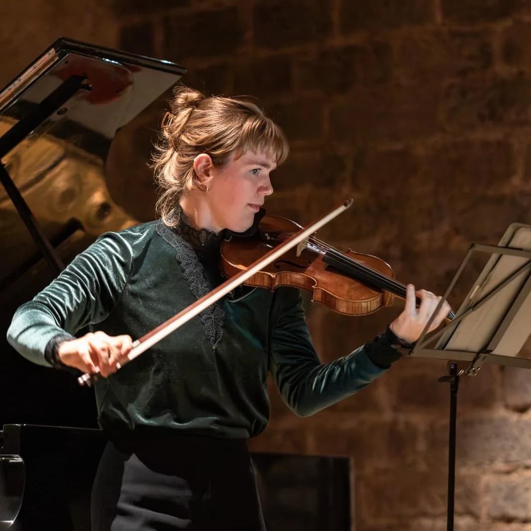 Annika Spegg spielt Geige im Profil in grünem Samtoberteil, im Hintergrund unscharf der Konzertflügel und das Notenpult eines weiteren Musikers.