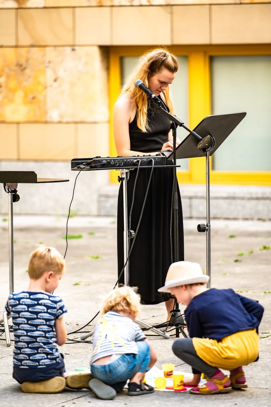Annika Spegg liest am Mikrofon hinter einem Keyboard im Außenbereich; vor ihr spielen drei kleine Kinder mit bunten Knetfiguren auf dem Boden, gelbe Fenster im Hintergrund.