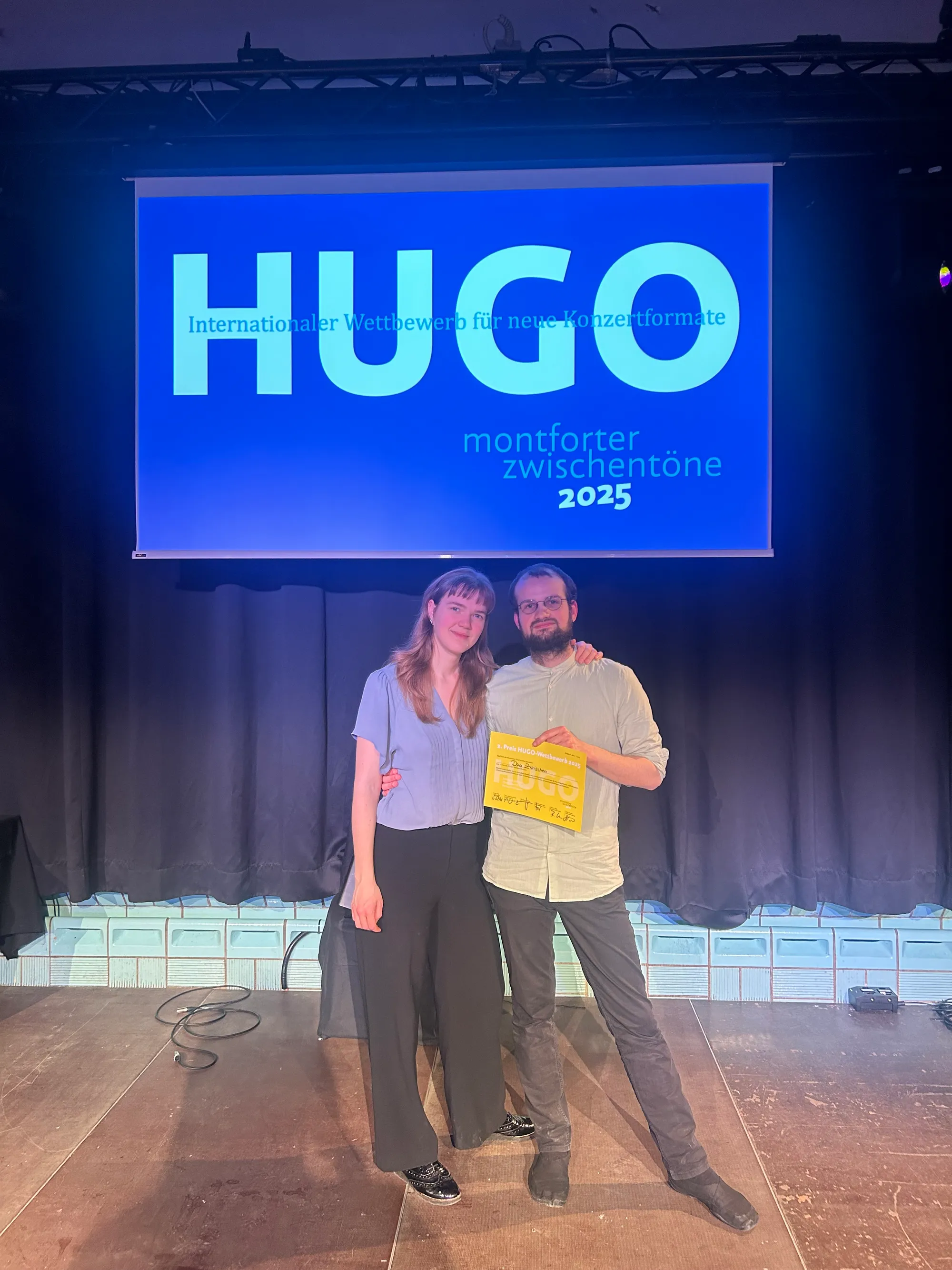 Annika Spegg und Robert Rülke (Duo Zwischen) auf der Bühne mit der gelben Urkunde des 2. Preises, im Hintergrund leuchtend blau das Logo „HUGO – Internationaler Wettbewerb für neue Konzertformate, Montforter Zwischentöne 2025“.