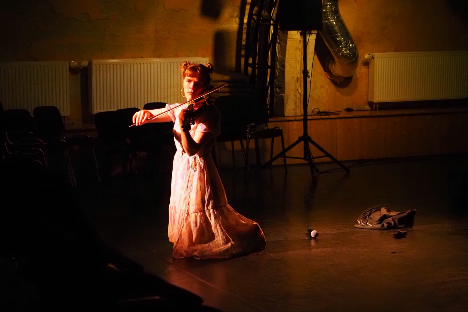 Szenenfoto Faust.Margarete — eine Performerin kniet im hellen Kleid und spielt Geige im warmen Spotlight.