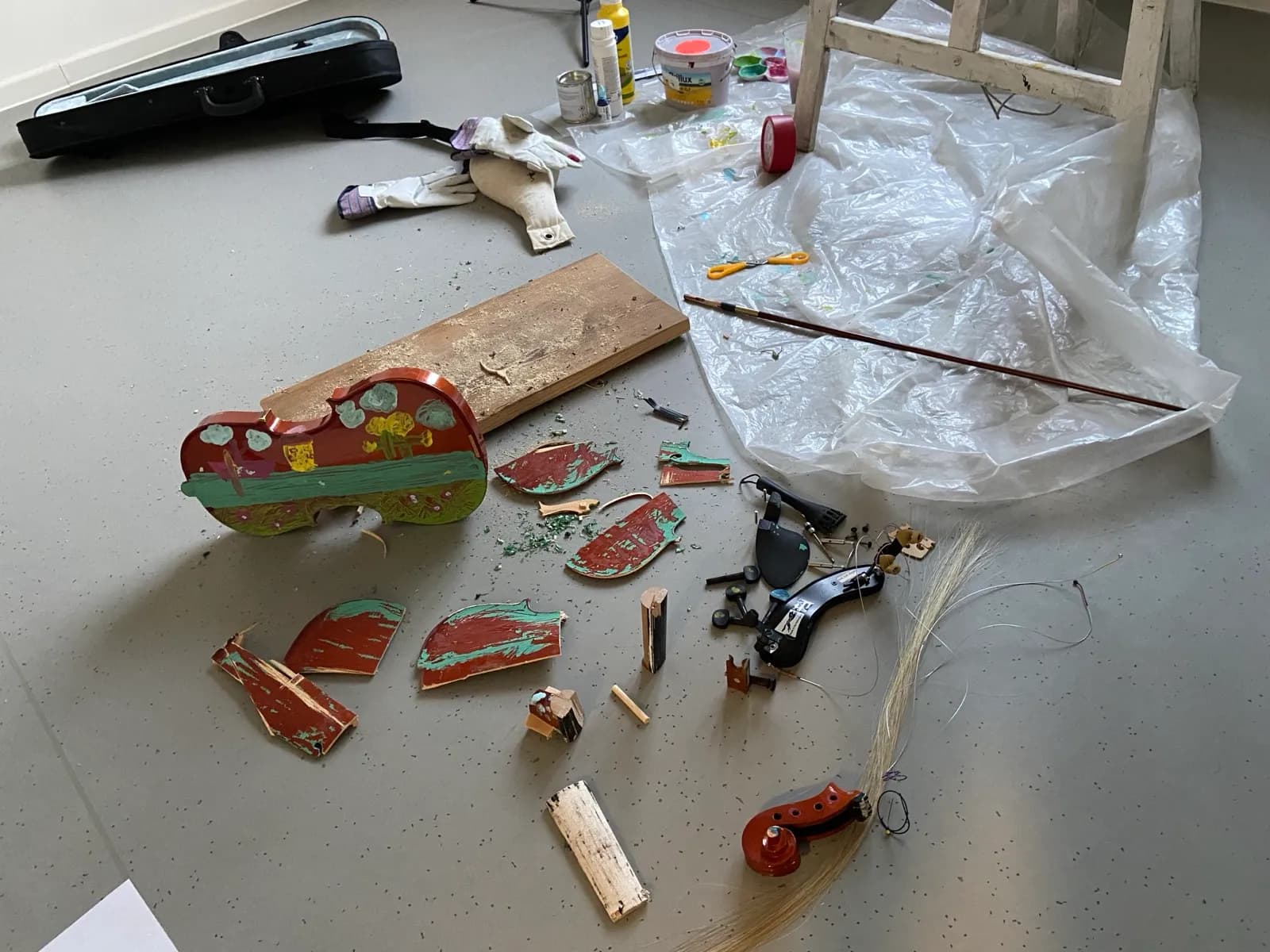 Werkstattszene: zerschlagene bemalte Geige in Einzelteilen auf dem Boden, daneben Geigenkasten, Bogen, Saiten und ein Schnittmuster-Handschuh — Spuren der Performance.
