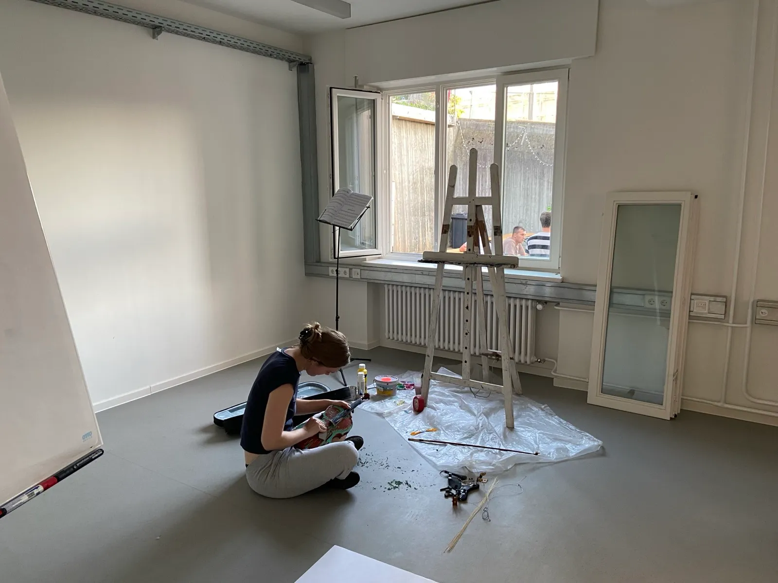 Annika Spegg sitzt im Schneidersitz im Probenraum auf dem Boden und arbeitet an der bemalten Geige; daneben die weiße Staffelei und ein Notenständer.