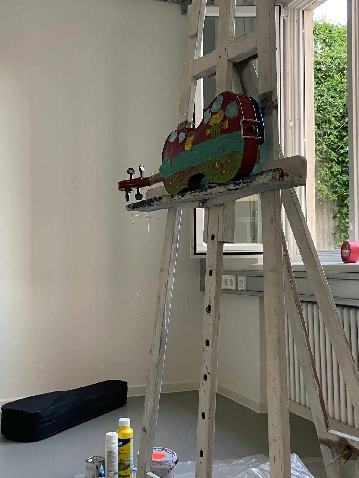 Bunt bemalte Geige (rot, türkis, grün, gelb) auf einer weißen Holzstaffelei vor einem Fenster mit Efeu im Hintergrund; daneben am Boden ein Geigenkasten und Acrylfarben.