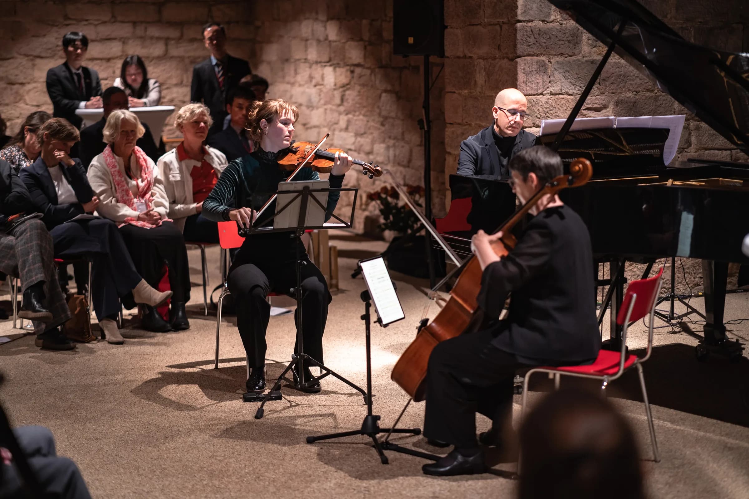 Klaviertrio im Konzert in einem Steinkellersaal: Annika Spegg an der Geige, Beverly Ellis am Cello und Alfonso Gomez am Flügel; vorne sitzendes Publikum, hinten stehende Gäste der koreanischen Delegation.