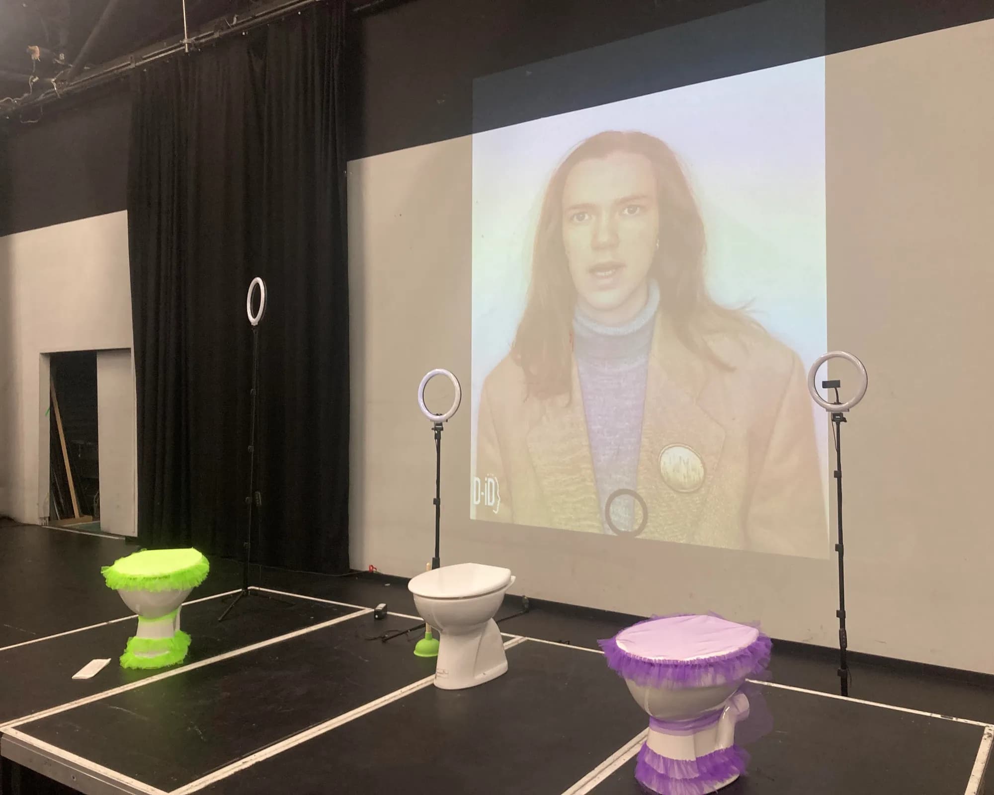 Bühnensituation KommentarTheater: drei Toiletten mit Tüllröcken in Neongrün, Weiß und Lila stehen vor zwei Ringlichtern, im Hintergrund Projektion eines AI-generierten sprechenden Kopfes (D-ID).