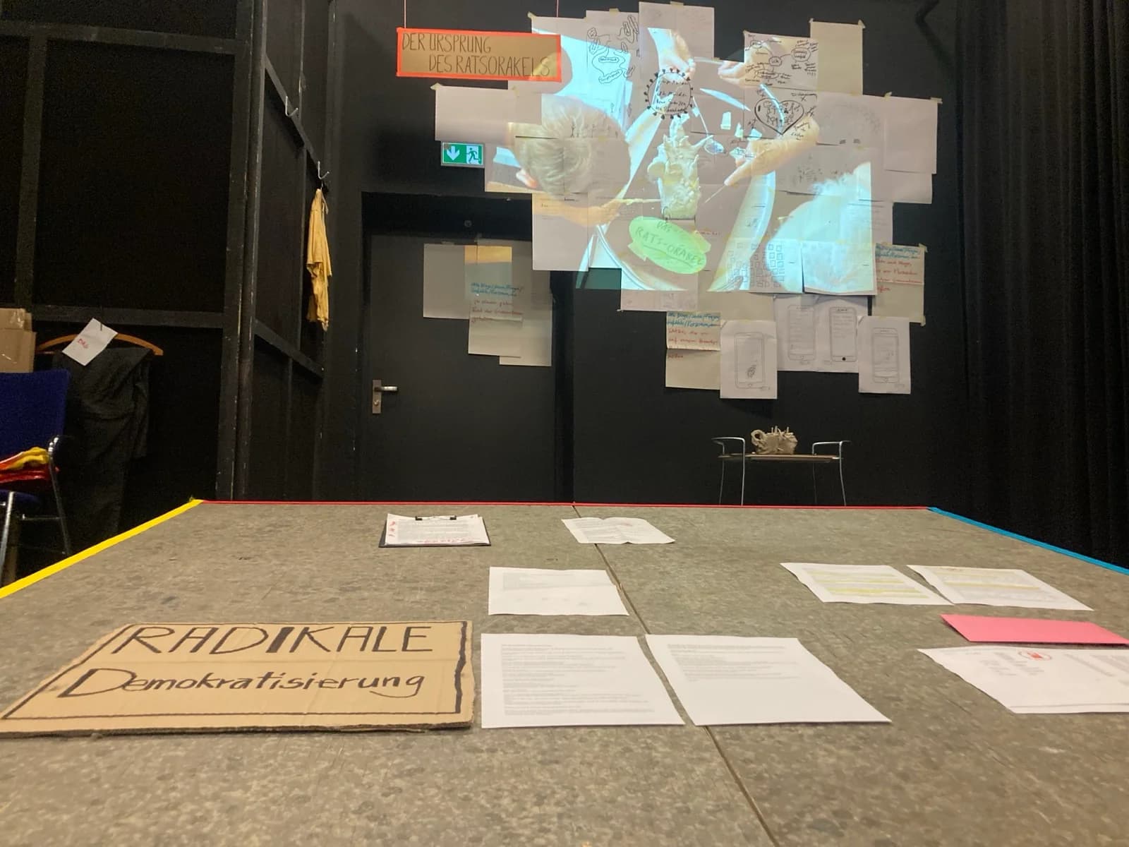 Recherche-Installation: Tisch mit Pappschild „RADIKALE Demokratisierung“, an der Wand Projektion „DER URSPRUNG DES RATSORAKELS“ über handbeschriebenen Papieren.