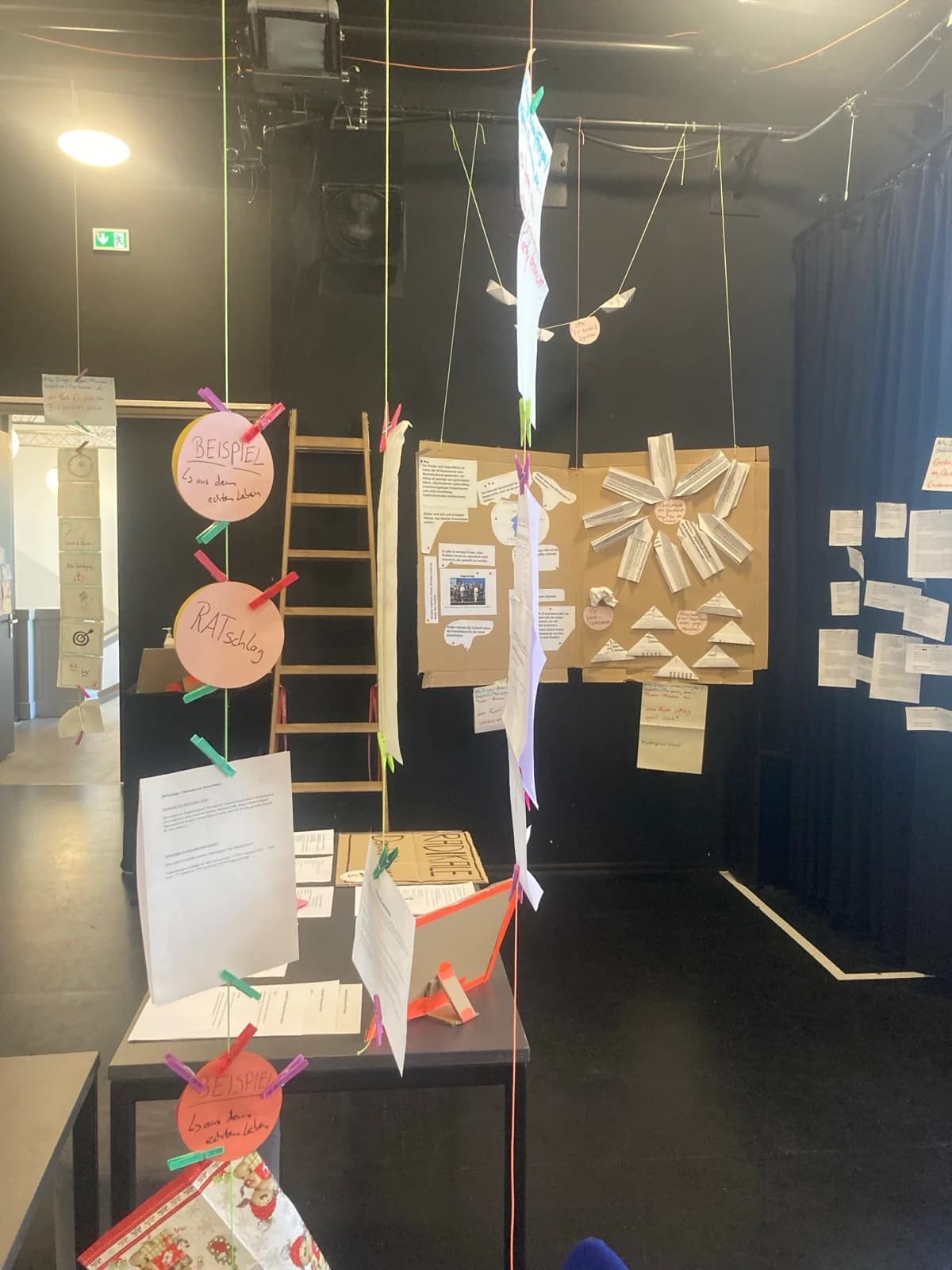 Hängende Papier-Installation mit den Karten „BEISPIEL aus dem echten Leben“ und „RATschlag“, dahinter eine Pappwand mit Notizen und Origami-Faltungen.