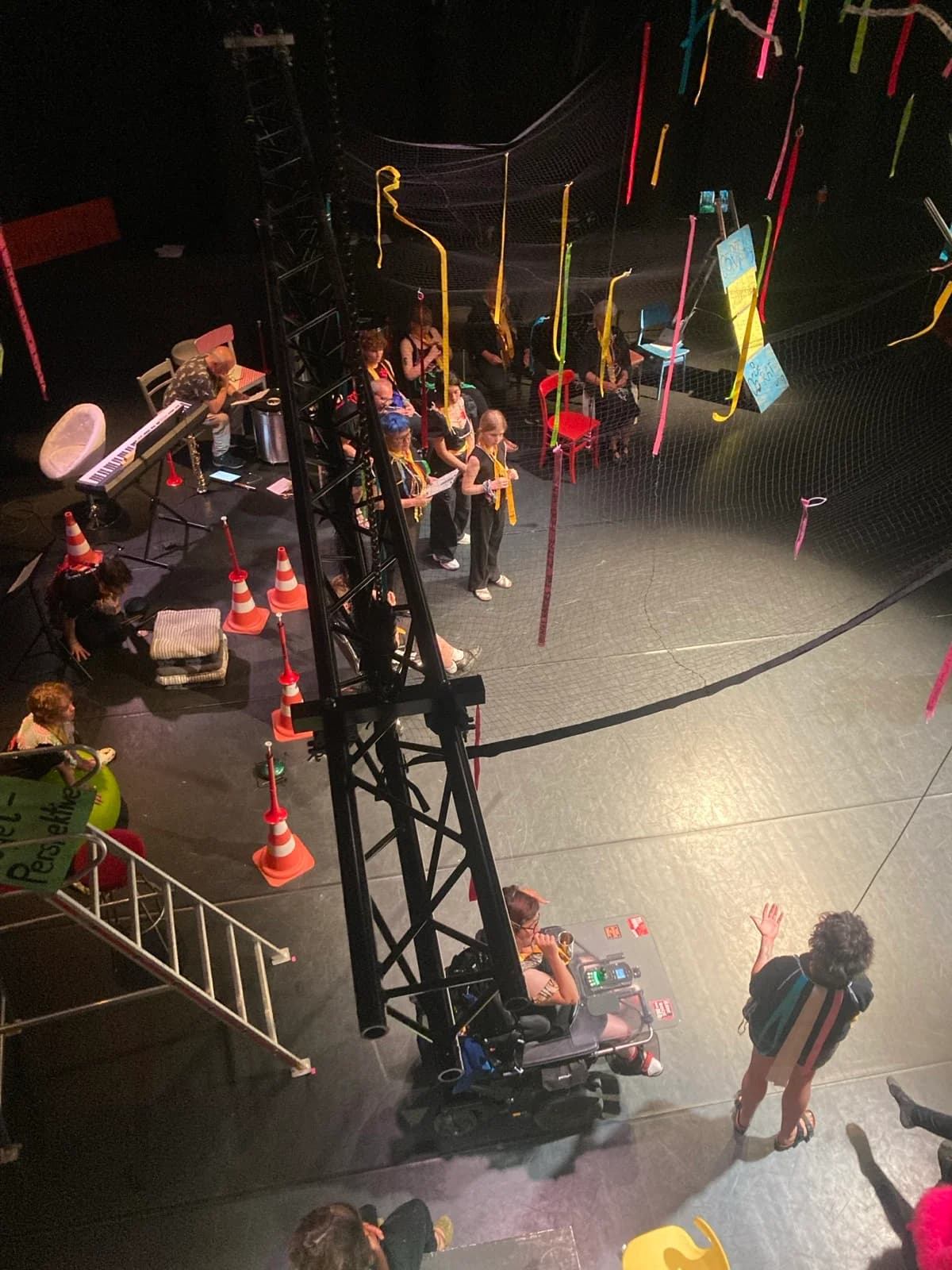 Rat-der-Generationen-Bühne im Theater der Jungen Welt: ein schwarzer Stahlmast mit aufgehängtem Netz, daran bunte Bänder, Pylonen und beschriftete Schilder; eine Person im Rollstuhl und Performende in der Mitte.