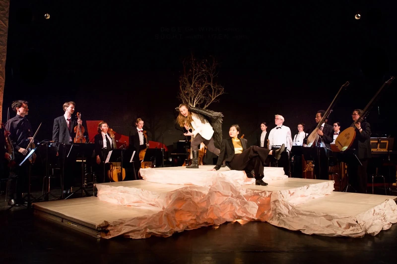 Bühnenbild Venus and Adonis: zerknülltes weißes Papier als Inselformation, ein Performer mit Flügeln und Sängerin in goldener Bluse darauf, im Hintergrund das Barockorchester mit Theorbe, Laute und Streichern.