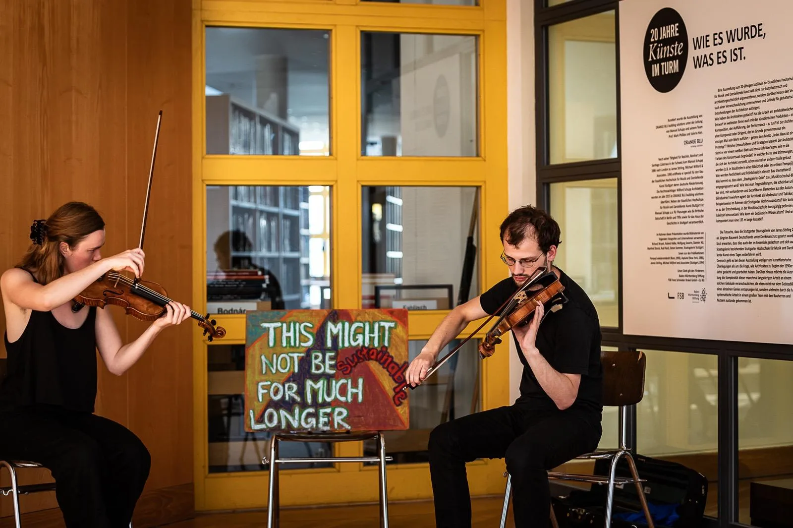 Duo Zwischen im Konzert in der Hochschulbibliothek; auf einem Notenständer ein Schild mit der Aufschrift 'This might not be sustainable for much longer'.