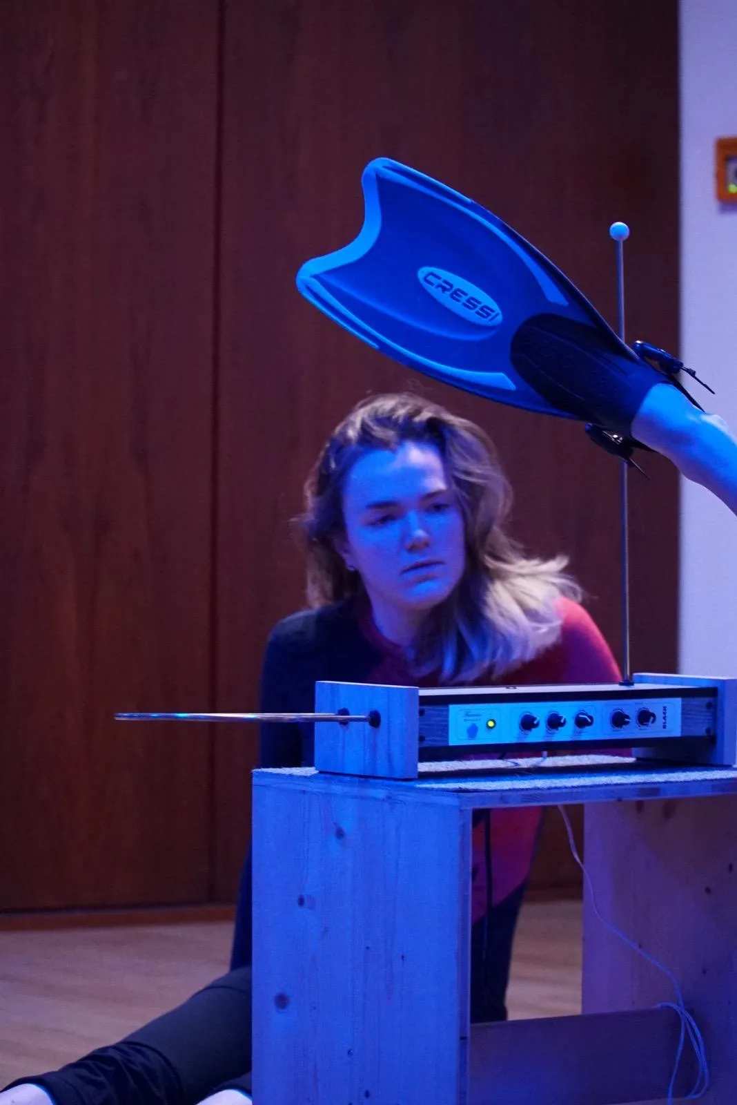 Annika Spegg sitzt am Boden mit elektrischer Klangapparatur; eine blaue Schwimmflosse wird über ihren Kopf gehalten.
