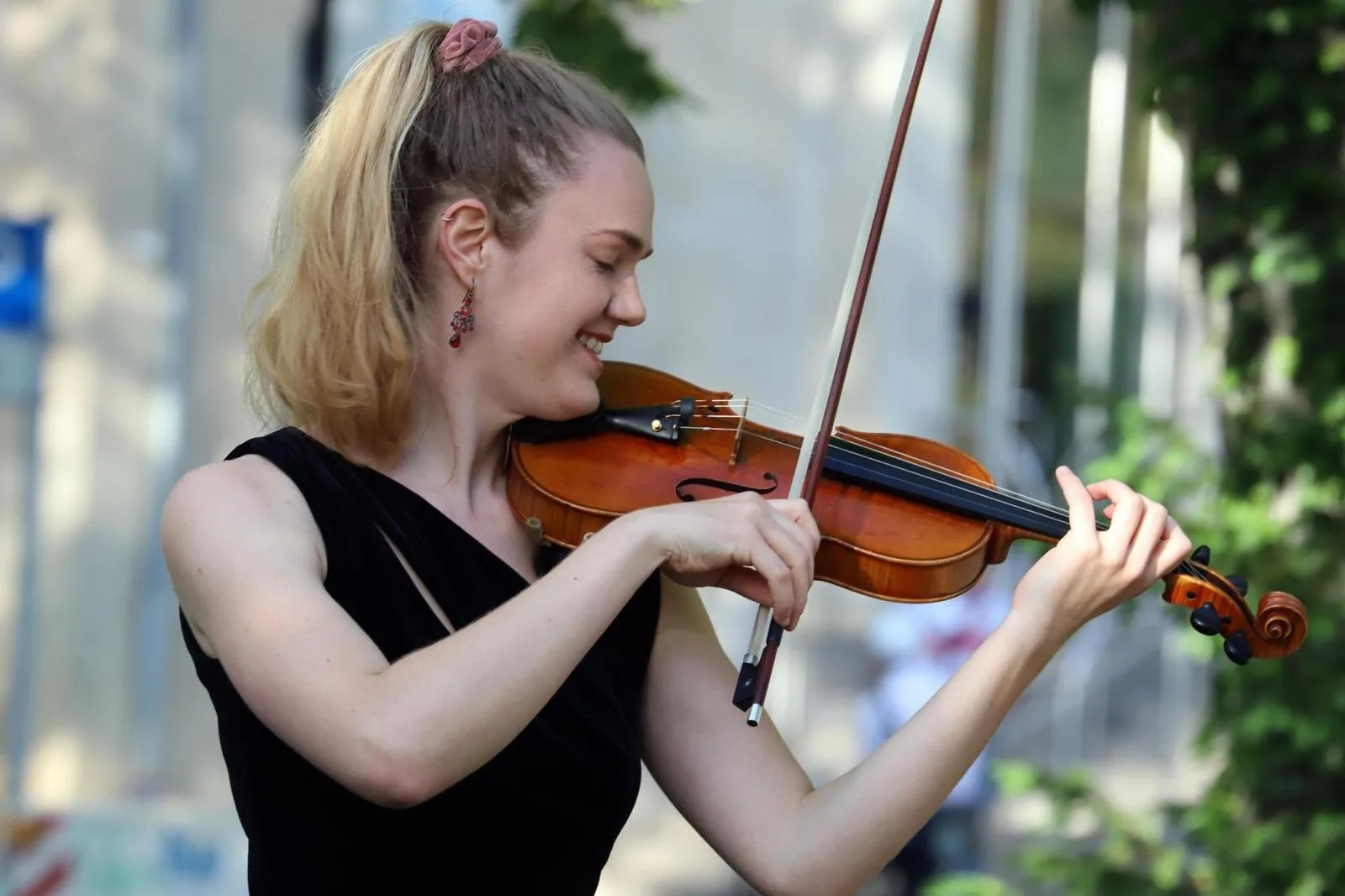 Annika Spegg lächelt beim Geige spielen im Sommer, im Profil vor unscharfem grünem Laub.