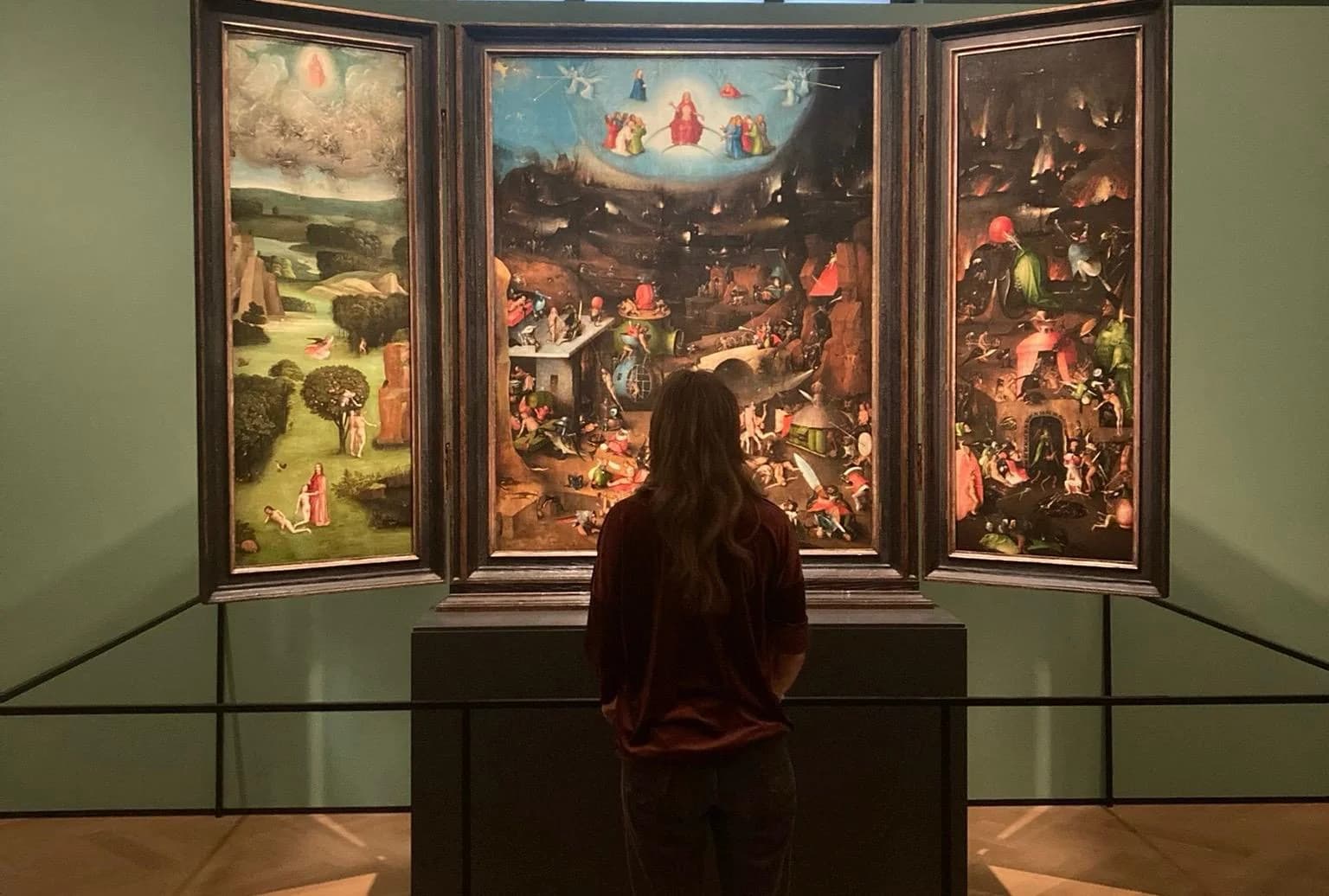 Annika Spegg von hinten vor einem Triptychon (Weltgericht-Altar) im Museum — Paradies, Endgericht und Hölle in dunklen Grün- und Erdtönen.